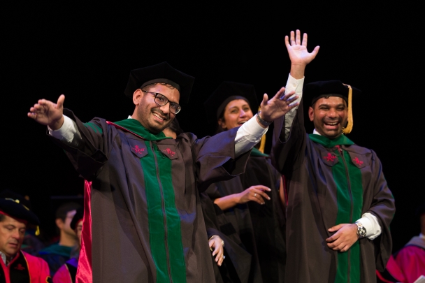 Graduates in black and green caps and gowns wave to the crowd in the theatre during convocation